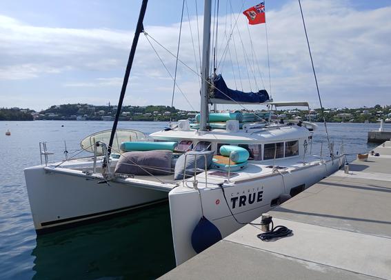 50-foot white sailing catamaran tied up on a float on a sunny day, flying a Bermuda flag.