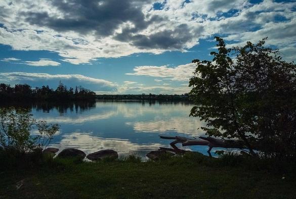 Looking across the calm waters of a lake with clouds reflecting in the water. The dark silhouette of trees and the distant shoreline are also seen.
