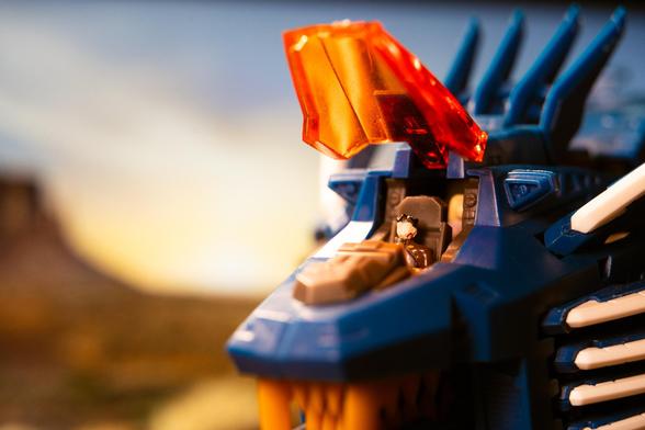 A close-up of a blue lion-like robot with an orange cockpit canopy, showing a small figure seated inside. The background features a blurred natural landscape.