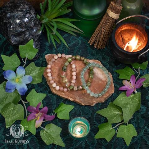 The three gemstone bracelets in our Litha Gemstone Bracelet Set are arranged on a slab of red stone. The elastic bracelets are made of round beads with different gemstones for each bracelet: sunstone, green lepidocrosite, and unakite. This is an exclusive product from Inked Goddess Creations.