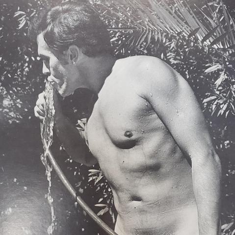 A cropped photo of an old black and white page from a male nudists magazine. A naked man, visible from the waist up, drinks water from a garden hose, outdoors, standing near fern-like shrubbery. The page is semigloss and there is reflective glare along the left, particularly in the lower half of the page. The glare does not obscure the subject matter.