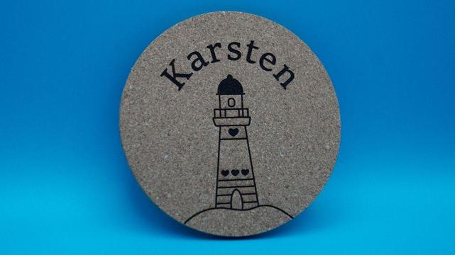 Runder Korkuntersetzer mit graviertem Leuchtturmmotiv und dem Namen „Karsten“ in gebogener Schrift darüber.