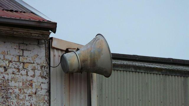 a photograph of a large rusty loudspeaker mounted on a corrugated metal wall. Photo attribution: Flickr user theenmoy