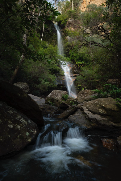 Waterfall coming over warm coloured sandstone cliffs surrounded by trees and in the foreground flowing towards the viewer over a small cascade. 