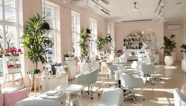 Bright nail salon interior with pastel colors, sunlight through large windows, manicure stations, and hands showing spring-themed pastel and bold red nails.