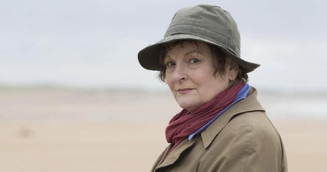 A woman with short brown hair wearing a green hat, a brown coat, and a red scarf stands on a beach, looking directly at the camera with a neutral expression. The background features a blurred sandy beach and cloudy sky. #DCIVeraStanhope