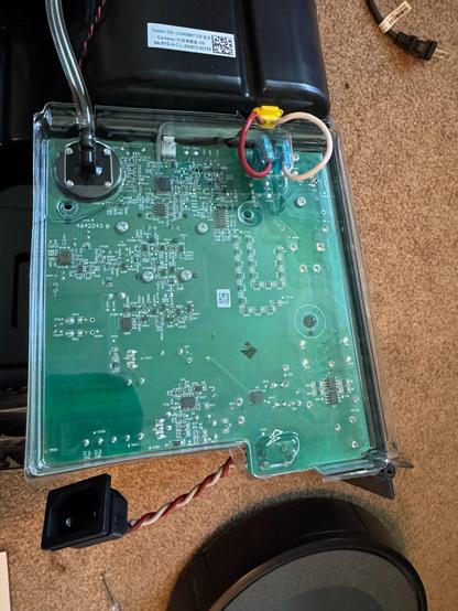 This image shows the inside of an electronic device, likely part of a control system. At the center is a green printed circuit board (PCB) with numerous tiny components and solder points. Several wires are connected to the board: a red-and-white twisted pair, a red wire, and a white wire, each leading to different parts of the device. The red and white wires are secured with a yellow adhesive near the top left. At the bottom, a clear plastic tube is attached to a black circular component, possibly a sensor or pump. The board is housed in a clear plastic cover, revealing intricate electronic pathways and parts.