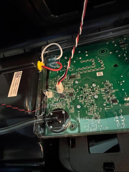 This image shows the inside of an electronic device, likely part of a control system. At the center is a green printed circuit board (PCB) with numerous tiny components and solder points. Several wires are connected to the board: a red-and-white twisted pair, a red wire, and a white wire, each leading to different parts of the device. The red and white wires are secured with a yellow adhesive near the top left. At the bottom, a clear plastic tube is attached to a black circular component, possibly a sensor or pump. The board is housed in a clear plastic cover, revealing intricate electronic pathways and parts.