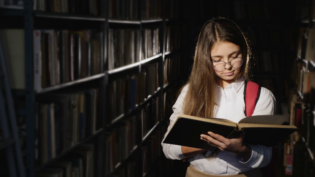 A girl in a dimly lit library wearing a red backpack and holding book, getting lost in its pages.