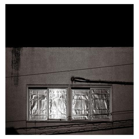 This monochrome image shows four side-by-side windows tightly sealed with reflective foil, crinkled and warped under tape. They sit in a plain concrete wall, bisected by deep shadows from overhead wires.