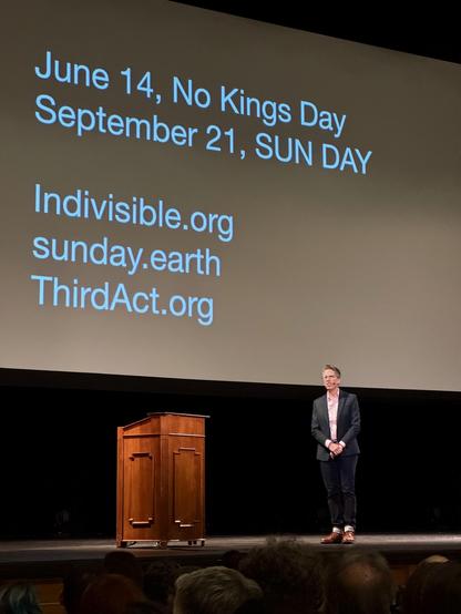 Alison Bechdel on stage at the Sydney Goldstein Theater. On the big screen behind her are a few dates of upcoming protests and some websites. The text says "Jun 14th, No Kings Day. September 21 Sun Day. Indivisible.org Sunday.earth ThirdAct.org". 