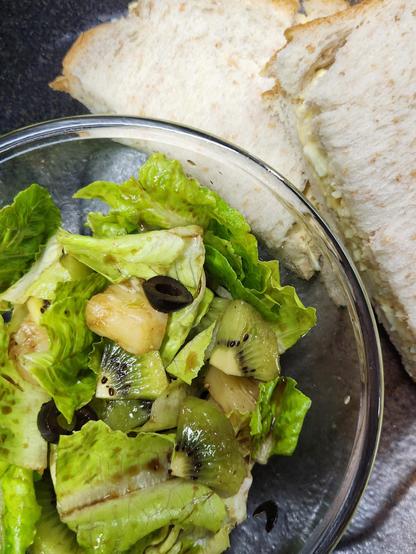 Fresh lettuce, pineapple chunks, black olives, and slices of kiwi in a clear glass bowl. The bowl is on a ceramic plate, beside a sliced egg sandwich.  
