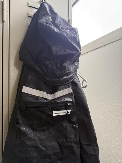 A wet black poncho hangs on a locker hook, featuring a reflective stripe and a zippered pocket. Natural light is coming from a nearby window, highlighting the water droplets on the fabric.