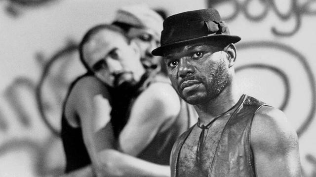Photo en noir et blanc montrant Riggs au premier plan, regard caméra, et deux hommes noirs enlacés en arrière plan, un peu flous.