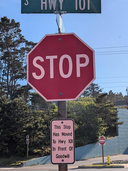 Picture of a stop sign on highway 101. Below the sign on the same post, a small, rectangular, white sign reads "This Stop Has Moved To Hwy 101 In Front Of Goodwill."