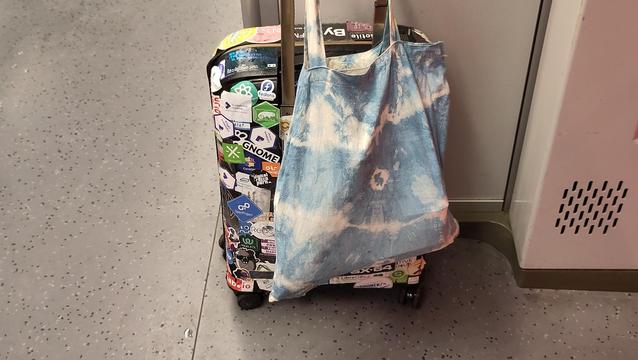 A trolley covered with open source stickers and a toat bag hanging over it. The trolley is standing in the corner of a regional train.