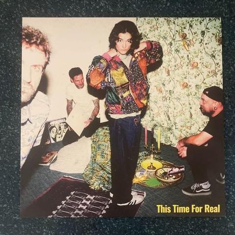 Eine Gruppe von vier Männern in einem farbenfrohen Innenbereich. Ein Mann steht in der Mitte und trägt eine leuchtende Jacke, während die anderen um ihn herum sitzen oder teilweise sichtbar sind. Im Hintergrund sind gemusterte Tapeten und ein Teppich zu sehen.