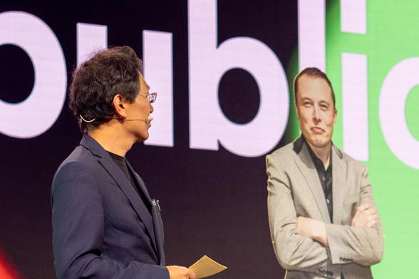 Bühne Stage 1. Anwalt Jun mit dem Kopf nach links gewandt, es sieht aus als ob er den Pappkamerad Musk direkt ansieht. Musk-PappKamerad schaut süffisant Richtung Jun. In Wahrheit schaut Jun zur Kollegin Jessika Flint (nicht im Bild).