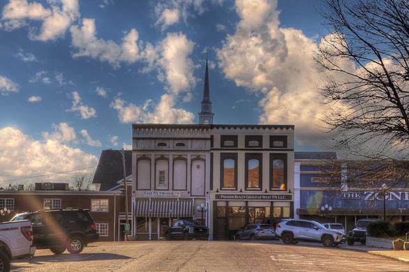 A small town square displays a row of historic buildings under a sky filled with fluffy clouds