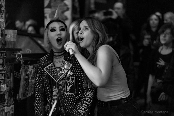 Two women are singing passionately on stage in a lively venue. One has a punk-inspired look with a spiked jacket and bold makeup, while the other is wearing a fitted top and holding a microphone. The background features an attentive crowd.