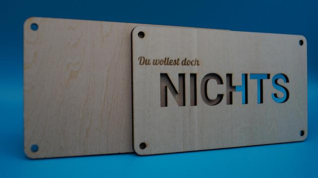 Holzplatte mit ausgeschnittener Aufschrift „Du wolltest doch NICHTS“, die Rückseite noch offen – vorbereitet als humorvolle Geschenkverpackung.