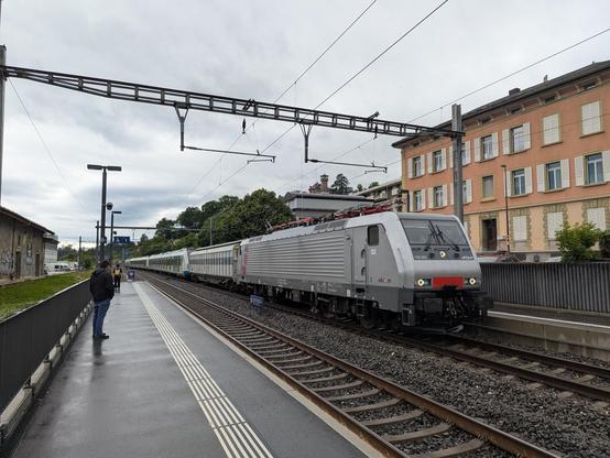 Une locomotive akiem de type traxx livrée grise inox tire deux S-Bahn de Stuttgart à travers la gare de Clarens sous un ciel pluvieux.
