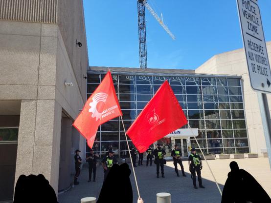 Three figures blacked out in the foreground. 11 police at the Ottawa police station on Elgin st in the background. Two red flags for the Communist Party of Canada are waving.