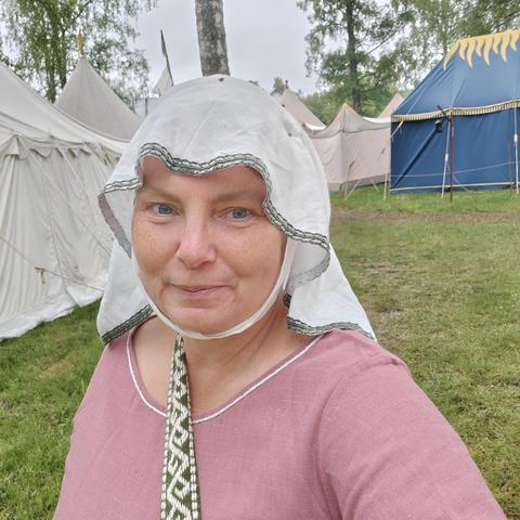 A smiling white middle aged woman in a pink dress and a white veil, with several medieval style tents behind .