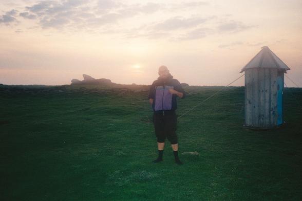 Un portrait de moi sur une île bretonne, au loin le soleil se couche sur les rocher et l'herbe verte.