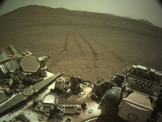 An ochre toned image capturing a Martian landscape, with the deck of the rover in the foreground, and two intertwined pairs of tracks receding away in the distance.