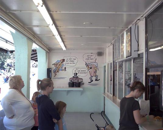 A snack bar with people lined up on the left, service windows to the right. On the cinder block wall in the back are painted three  anthropormorphic food items: a hot dog dressed as a cowboy, a cup of soda with a grey goatee, and a hamburger in a top hat. Respectively, they're saying, "I'm old time Hot Dawg good anytime", "Git me! Old Soda Pop!", and "Hi! I'm Ham-Burger I'm available!"
