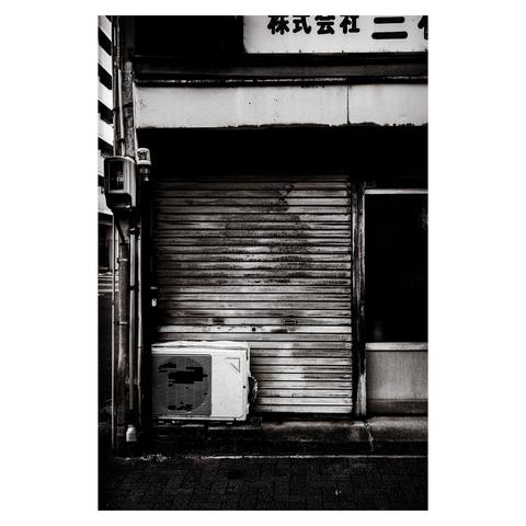 This monochrome photo captures a rust-streaked metal shutter with an aging air conditioning unit hunched below it, missing several grille slats. The scene is perfectly quiet—no signage suggesting what kind of business once lived behind the shutter, just a tired facade and an appliance long past its prime.