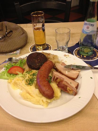 The image depicts a meal served on a white plate, placed on a wooden table. The plate contains a variety of food items, including a sausage, slices of bacon, mashed potatoes, a slice of black pudding, sauerkraut, and a slice of tomato garnished with lettuce and chives. A fork is positioned to the left of the plate, and a knife is on the right. In the background, there is a glass of beer with a golden color, a clear glass, and a bottle of Gerolsteiner Sprudel water. A hat and a pair of glasses a…