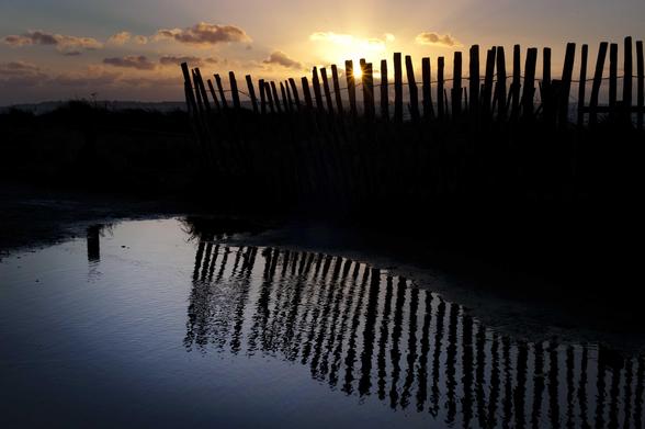 L’image est séparée en trois: en bas une grande flaque d’eau, au centre une bande sombre avec une clôture en bois et en haut, le ciel. La clôture que l’on voit en ombre chinoise (contre-jour) se reflète dans la flaque. Entre deux piquets de la clôture, le soleil se couche et éclaire les rares nuages.