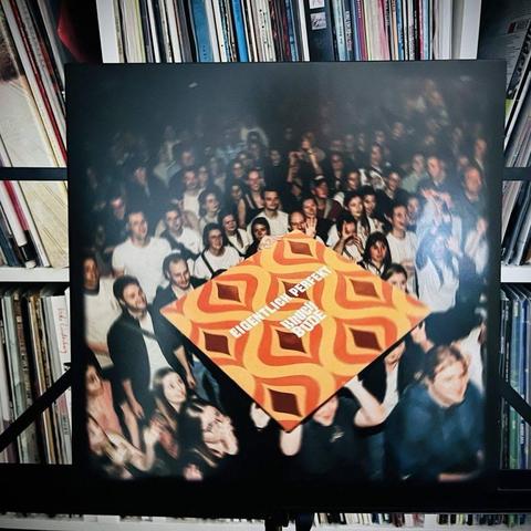 Eine Schallplattenhülle mit einem abstrakten Muster in Orange und Braun wird vor einer Menschenmenge auf einer lebhaften Veranstaltung ausgestellt, während im Hintergrund ein mit Schallplatten gefülltes Bücherregal steht.