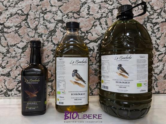 Tres botellas de aceite de oliva virgen extra ecológico "La Bandada" de diferentes tamaños se muestran sobre una superficie de granito. La botella más pequeña contiene 1 litro, la botella mediana 2 litros y la más grande 5 litros. Todas las botellas muestran una imagen de un pájaro y la etiqueta indica que el aceite es ecológico y orgánico. El texto "Biolibere, supermercado cooperativo" aparece en la parte inferior de la imagen.