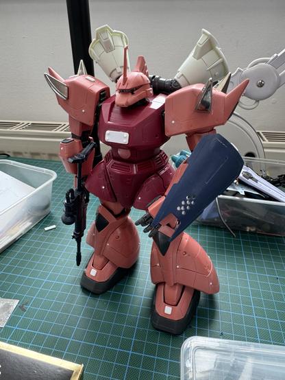 A large, articulated robot model stands on a textured green cutting mat. The robot is predominantly in shades of red and features armor plating, spikes, and a weapon in one hand. Surrounding the robot are various modeling tools and containers.