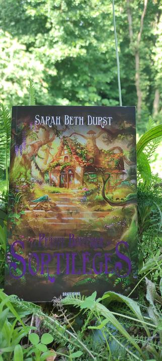 Livre intitulé La petite boutique de sortilèges de Sarah Beth Durst, posé dans un sous-bois luxuriant. Sa couverture colorée montre une maison de conte de fées, couverte de végétation et baignée de lumière dorée, avec un petit dragon ailé sur les marches. Le livre semble faire partie intégrante du décor naturel, entouré de fougères et de plantes sauvages sous une lumière d'été filtrée par les arbres