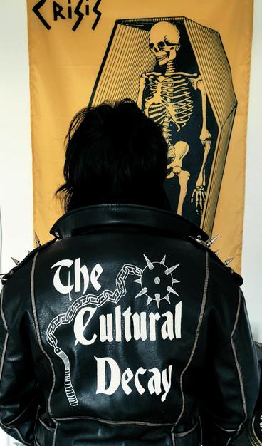 Una persona de espaldas con una chaqueta de cuero que tiene un diseño de "Cultural Decay" banda de Postpunk belga de culto de los 80's, y un fondo amarillo con una bandera de "Crisis". El diseño de la chaqueta tiene también un mangual, arma medieval con un mango hecho de madera del cual se desprende una cadena y que en la punta tiene una bola de metal con pinchos.