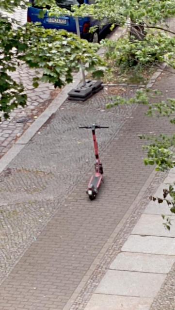 Das Bild zeigt eine rote Elektro-Scooter auf einem gepflasterten Radweg! Der Scooter steht aufrecht, mit dem Lenker nach oben und der Plattform nach unten. Der Gehweg besteht aus roten Steinen und ist von einem breiteren, grauen Gehweg auf der rechten Seite und einem gepflasterten Bereich auf der linken Seite abgegrenzt. Im Hintergrund ist ein blauer PKW zu sehen. Links im Bild sind grüne Blätter von Bäumen zu erkennen, die den Gehweg teilweise verdecken. Der Scooter ist in der Mitte des Bildes…