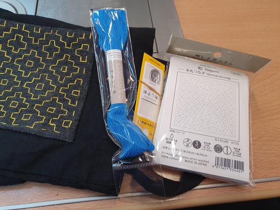 Ein in durchsichtiges Plastik verpacktes weißes Tuch , ein Päckchen Sticknadeln in gelber Papierverpackung und ein strahlend blauer Strang Sashikogarn liegen zusammen mit einer schwarzen Stofftasche mit gelber Stickerei auf einem Tisch im ICE. 
