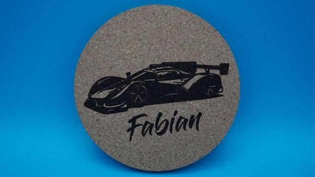 Runder Korkuntersetzer mit einem gravierten Rennwagen und dem Namen „Fabian“, vor blauem Hintergrund fotografiert.