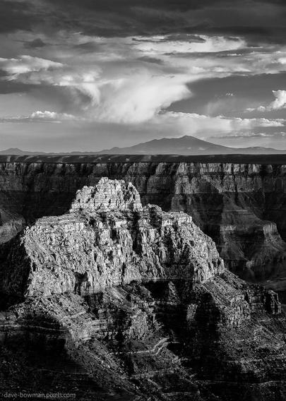 A photograph of the North Rim of the Grand Canyon in Arizona, captured in monochrome under late afternoon light. Rugged cliffs stretch across the frame, their jagged edges highlighted by deep shadows that accentuate the vast depth of the canyon. Above, dramatic clouds sweep through the sky, adding movement and contrast to the stark, expansive landscape.