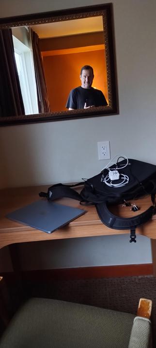 Photo of me looking into a mirror with laptop, bags, and charging cables on the desk