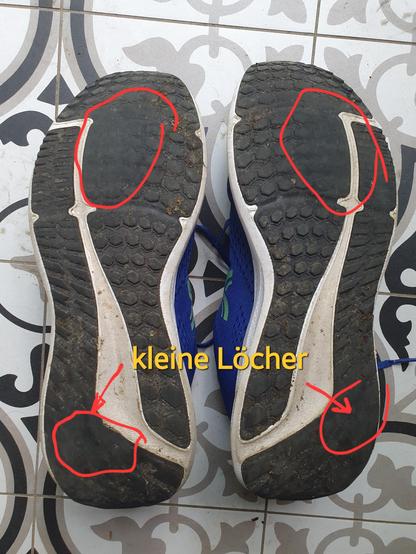 Das Bild zeigt die Sohlen von zwei Sportschuhen, die auf einem fliesenbepflasterten Boden liegen. Die Sohlen sind schwarz mit einem weißen Rand und haben verschiedene kleine Löcher, die mit roten Kreisen und Pfeilen markiert sind. Die Schuhe sind blau mit grünen Akzenten. Der Text "kleine Löcher" in gelber Farbe befindet sich in der Mitte des Bildes und deutet auf die beschädigten Bereiche hin. Die Schuhe scheinen abgenutzt zu sein, was durch die Löcher und die allgemeine Beschädigung der Sohle…