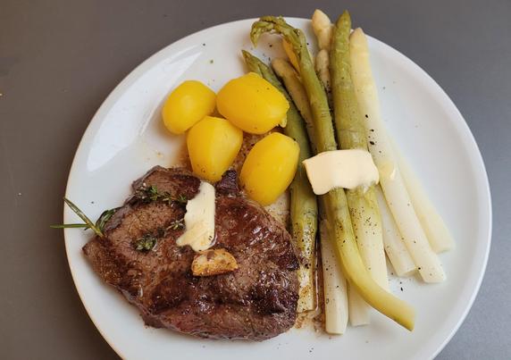 Farbfoto: Auf einem weißen Teller liegt das beschriebene Gericht, das Steak mit einer gebratenen Knoblauchzehe und Kräuterzweigchen, außerdem etwas Butter auf Fleisch und Spargel. 
