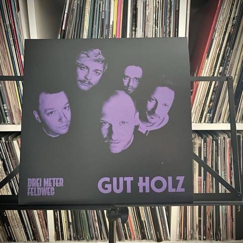 Ein Albumcover, das fünf Männer mit einem lilafarbenen, monochromen Design zeigt. Der Text "DREI METER FELDWEG" und "GUT HOLZ" ist am unteren Rand zu lesen. Der Hintergrund besteht aus gestapelten Schallplatten.