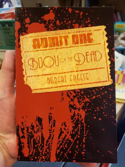 A photo of a softcover book cover. The book in my hand is "Bijou of the Dead" by Robert Freese. The black cover is printed with blood splatter and features a torn movie ticket stub with the title and author on it's face and "Admit One" printed across the top. In the edge  background of the photo the are other books and shelves visible but blurry.