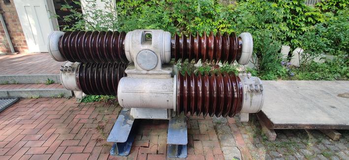 ein Hochspannungskondensator. stehen auf zwei doppel T Trägern.  ein metallischer Grundkörper an dem links und rechts jeweils zwei keramische Isolatoren angebracht sind. diese Elaine in einen Art dunkelrot gefärbt.