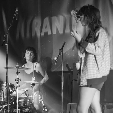 A black and white image of two female musicians performing on stage. One is playing the drums, while the other is playing the guitar. The background features a banner with the word "Niranium" and subtle lighting effects.